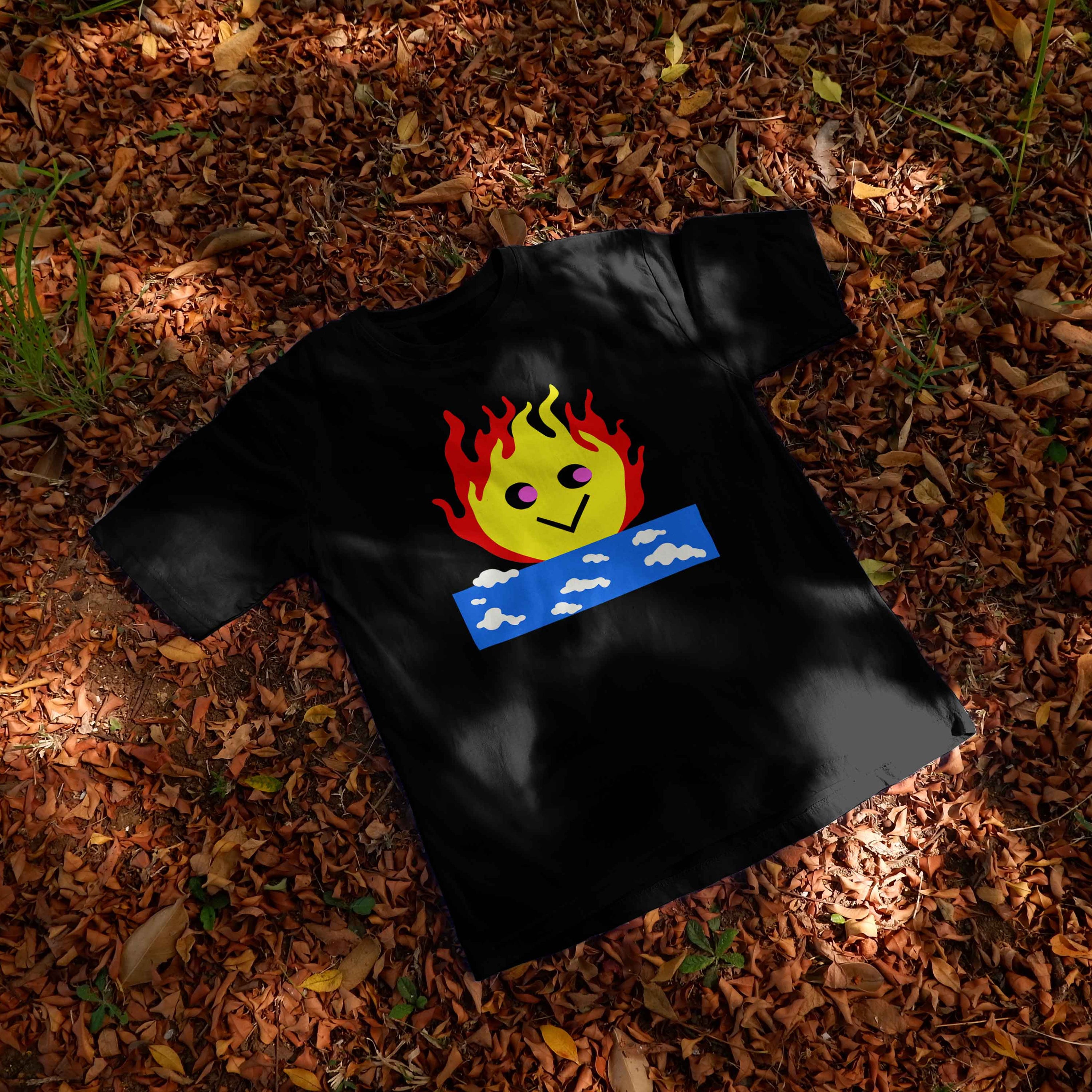 Black t-shirt with a design of smiley fire on top of a sky on a ground covered with dry leaves